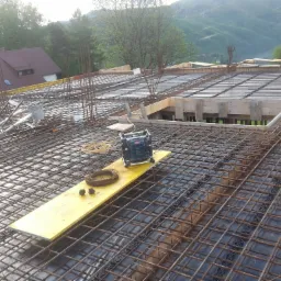 Zbrojenie płyty stropowej z widocznym deskowaniem, na tle górskiego krajobrazu i zabudowań.