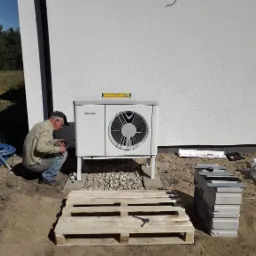 Instalator w trakcie montażu zewnętrznej jednostki pompy ciepła marki Stiebel Eltron, poziomica na obudowie, paleta i bloczki obok, widok na białą elewację budynku i las w tle.