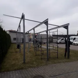 Montaż stalowej konstrukcji z profili na trawniku, widoczni pracownicy i drabina, w tle budynek i zaparkowane samochody dostawcze.