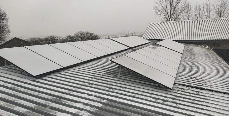 Panele słoneczne pokryte śniegiem na blaszanym dachu budynku gospodarczego w zimowy dzień.