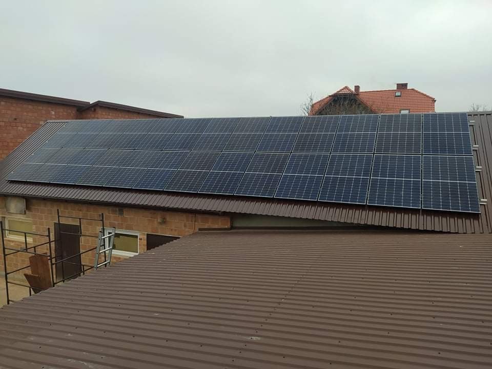 Panele słoneczne na brązowym blaszanym dachu budynku gospodarczego z czerwonej cegły, widok z innego dachu, pochmurne niebo.