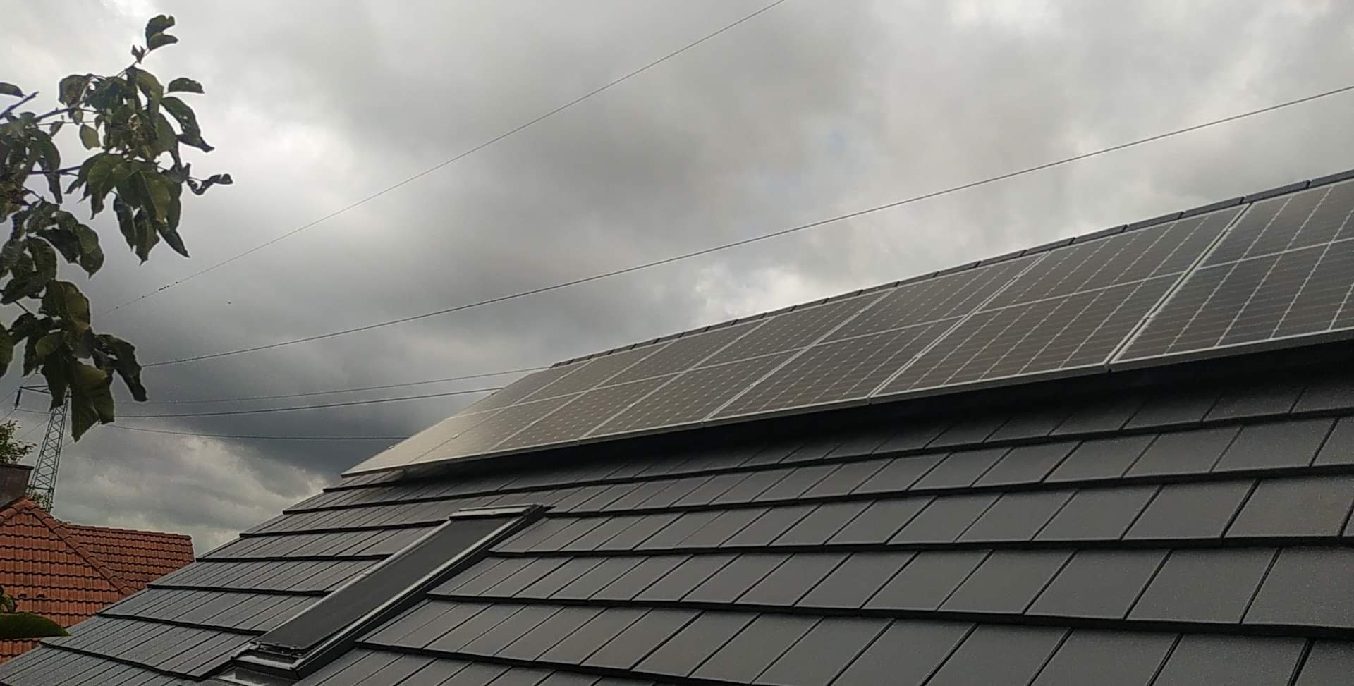 Ciemnoszare panele fotowoltaiczne zamontowane na dachu z dachówki bitumicznej w pochmurny dzień, widoczne linie energetyczne i fragment drzewa w lewym górnym rogu.