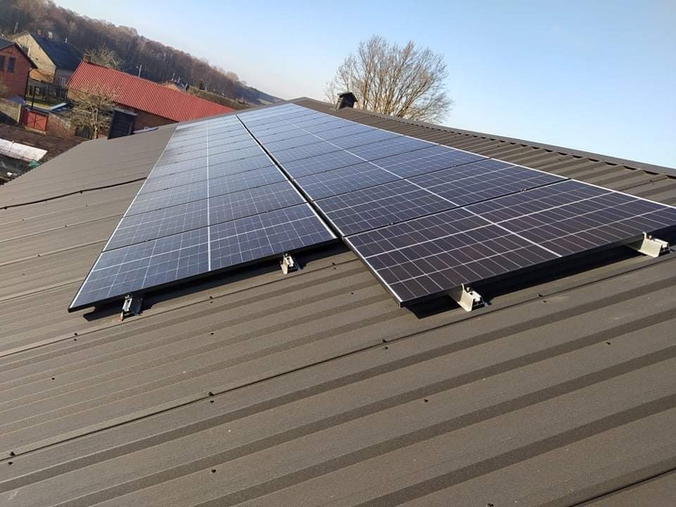 Panele słoneczne zamontowane na metalowym dachu z widocznymi mocowaniami i fragmentem zabudowań w tle, sfotografowane w słoneczny dzień.