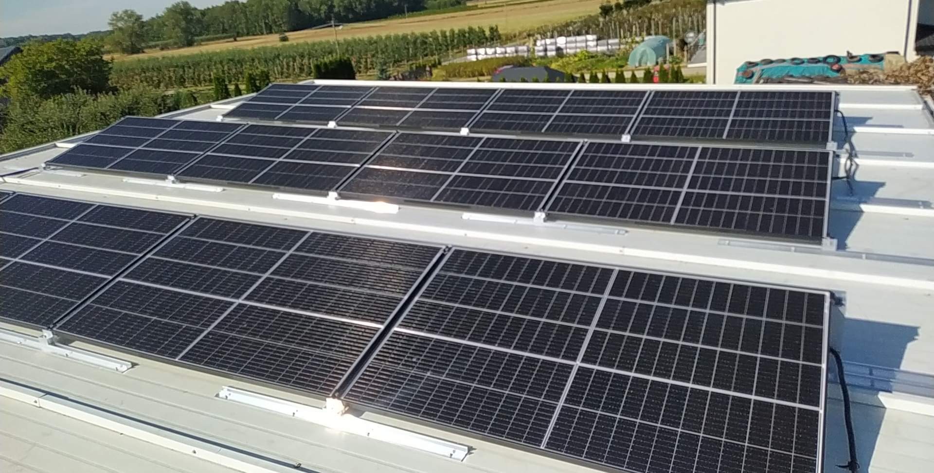 Panel słoneczny na jasnoszarym dachu płaskim z widocznymi elementami montażowymi, w tle zielony krajobraz z drzewami i zabudowaniami.