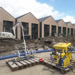 Zgrzewarka do rur z tworzyw sztucznych na palecie, na tle wykopu, minikoparki i szeregu budynków w trakcie budowy.