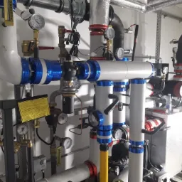 Rozbudowany system rur i zaworów w kotłowni, rury owinięte niebieską folią, liczne manometry i sterowniki, widoczny fragment pompy obiegowej i zbiornika wyrównawczego.