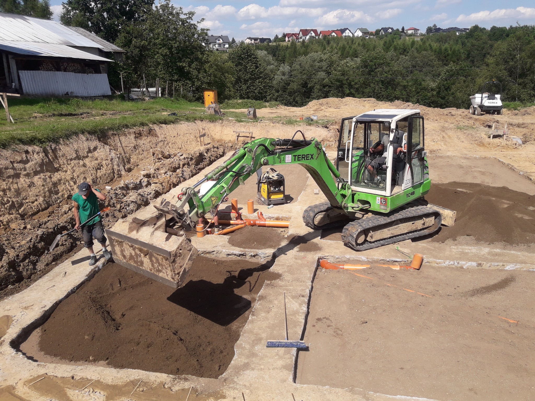 Prace ziemne: Wykop pod fundamenty domu jednorodzinnego z użyciem minikoparki Terex TC48, widoczne szalunki, rury kanalizacyjne, zagęszczarka i pracownik z łopatą. W tle zabudowa jednorodzinna.