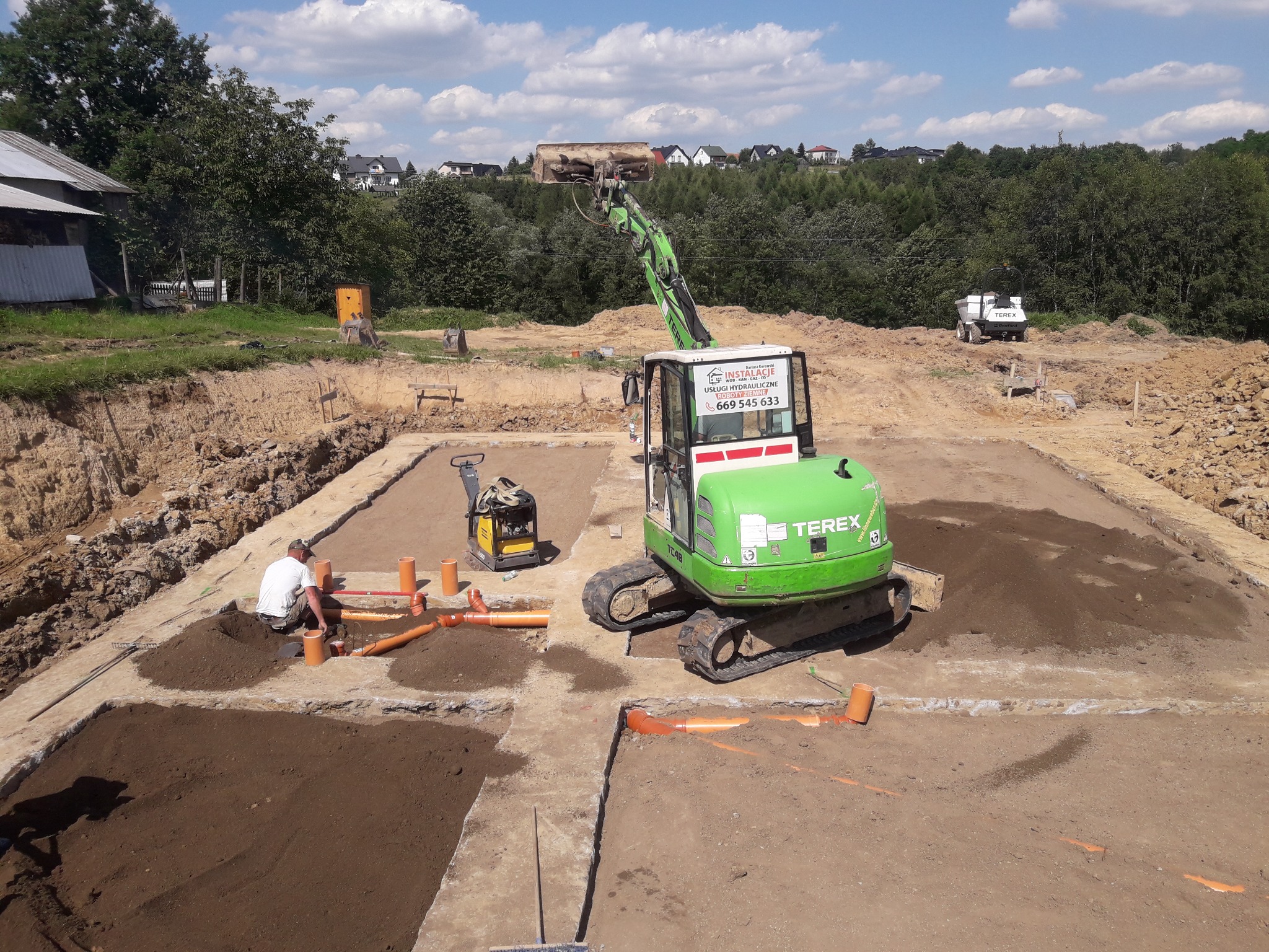 Fundamenty domu jednorodzinnego w trakcie budowy, widoczne wykopy z ułożonymi rurami kanalizacyjnymi, zagęszczarka wibracyjna i zielona mini koparka Terex z operatorem, w tle zabudowa jednorodzinna.