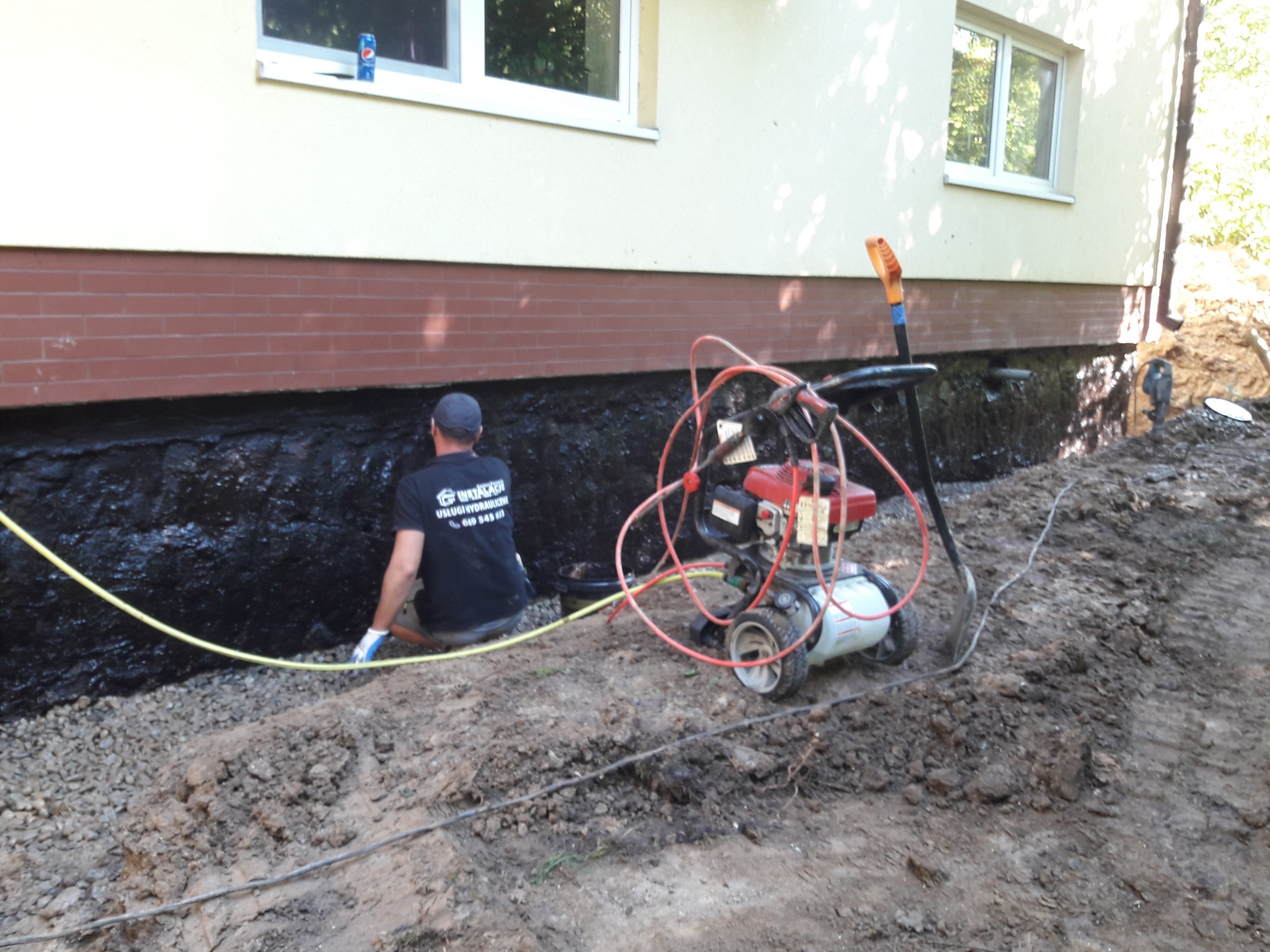 Pracownik w trakcie hydroizolacji fundamentów budynku, widoczna czarna powłoka izolacyjna, agregat malarski, wykop, w tle puszka Pepsi na parapecie okna.