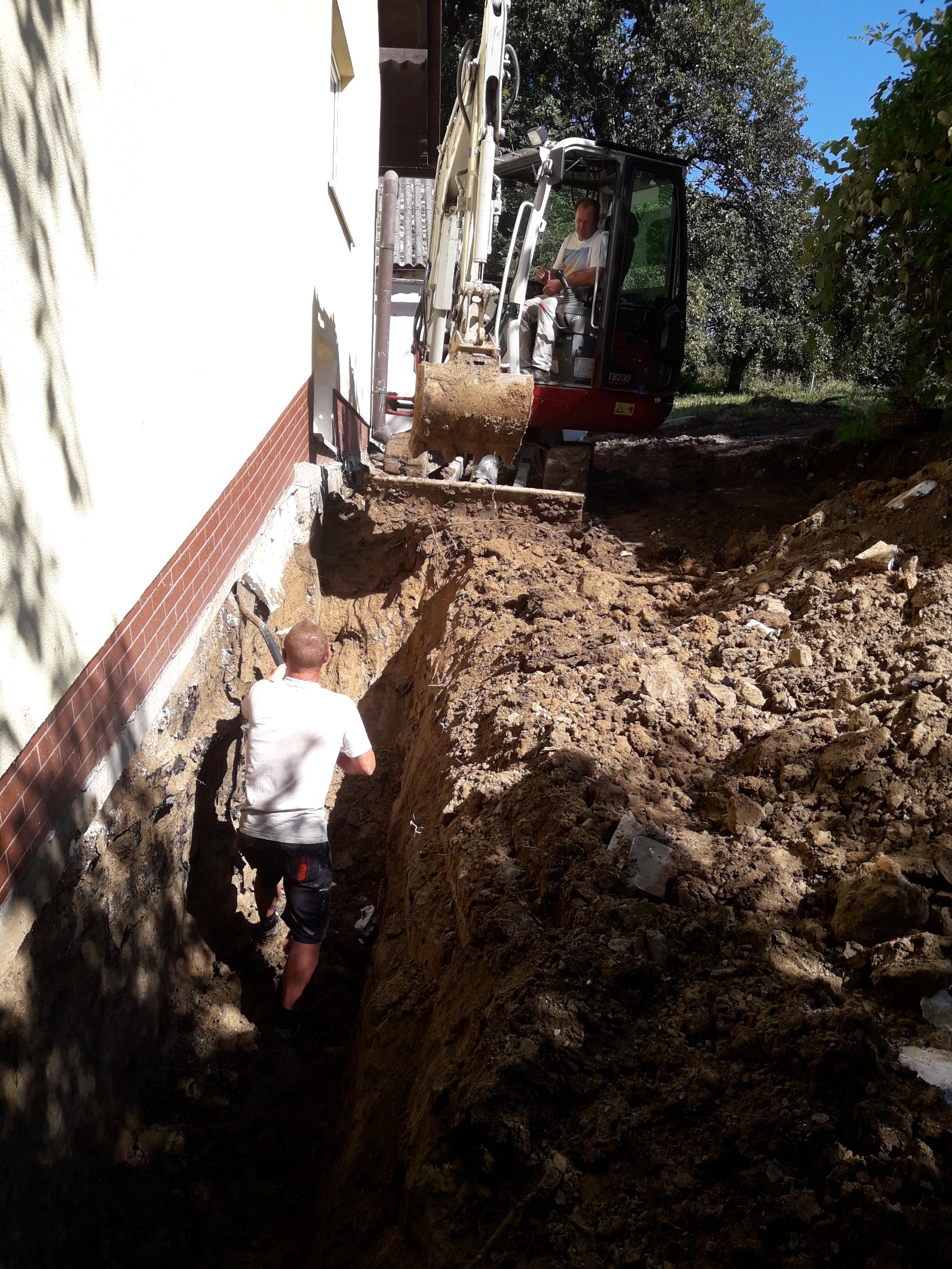 Wykop pod fundamenty domu jednorodzinnego wykonywany koparką TB230, z pracownikiem w wykopie wyrównującym ściany szpadlem.