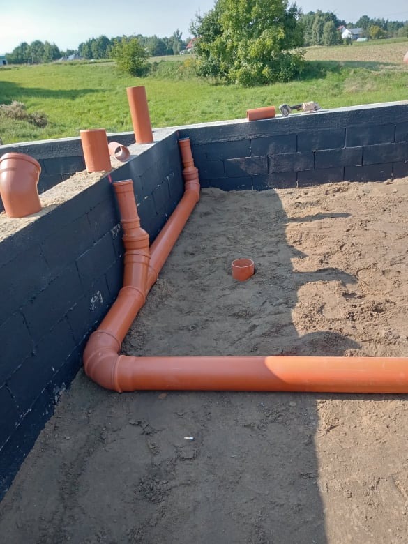 Fundamenty z izolacją i częściowo ułożonym systemem rur kanalizacyjnych z tworzywa sztucznego, widok z góry na plac budowy w słoneczny dzień.