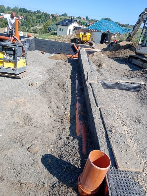 Budowa fundamentów domu jednorodzinnego z widocznymi rurami kanalizacyjnymi, ubijarką oraz koparką w tle.