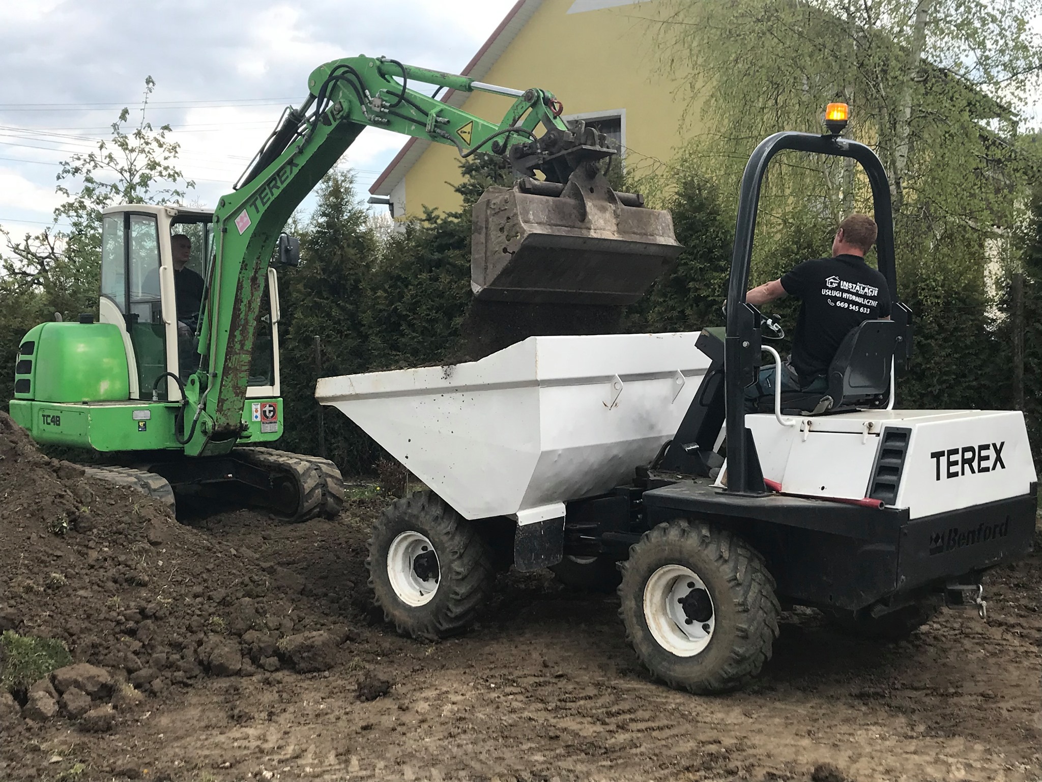 Zielona koparka Terex TC48 nabiera ziemię do białej wywrotki Benford, operator w czarnej koszulce z logo hydraulicznym, przygotowanie terenu pod budowę.