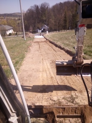 Wykop pod instalację, widok z kabiny koparki, widoczne ślady gąsienic i rów zabezpieczony białą folią, w tle budynki mieszkalne.