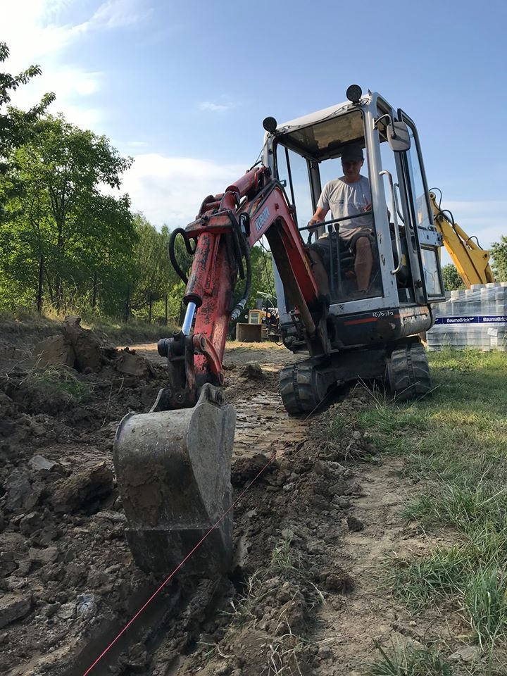 Operator koparki Kubota w trakcie prac ziemnych, wykop pod instalacje, widoczna wytyczona linia, w tle drzewa i palety z materiałami budowlanymi.
