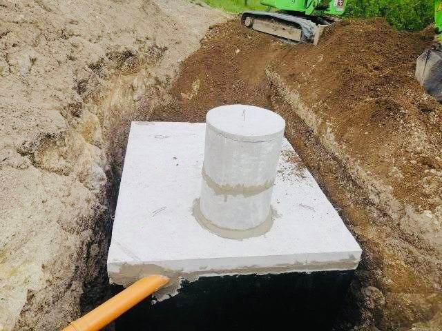 Betonowy zbiornik z pokrywą, częściowo zakopany w ziemi, widoczny pomarańczowy przewód odpływowy i koparka w tle.