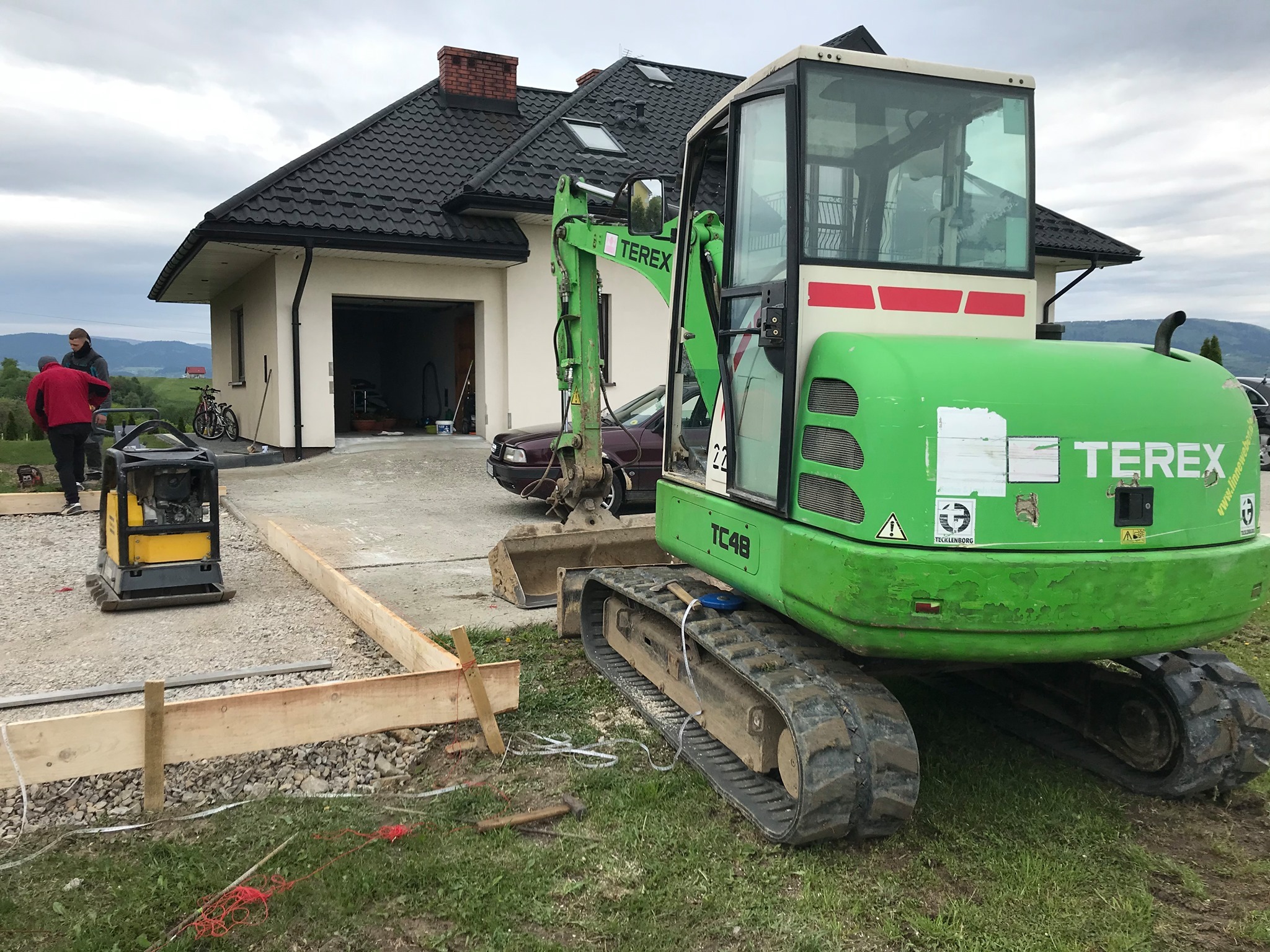 Zielona minikoparka Terex TC48 na placu budowy, widoczne drewniane szalunki, ubijarka, dom w tle i pracownicy, teren przygotowywany pod wylewkę betonową.