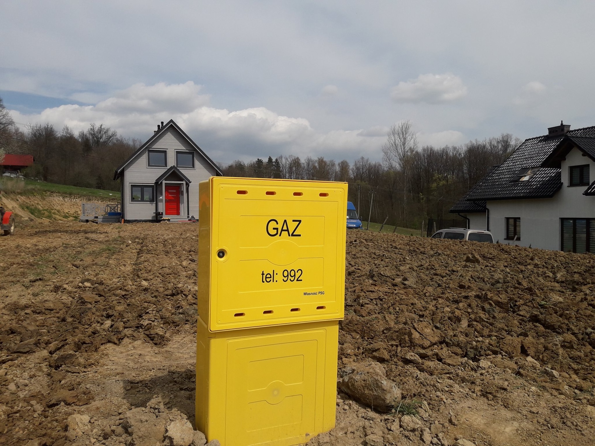 Żółta skrzynka gazowa z napisem 'GAZ' i numerem alarmowym 992 na tle świeżo zaoranej ziemi i dwóch domów w różnym stylu architektonicznym, częściowo pochmurne niebo.