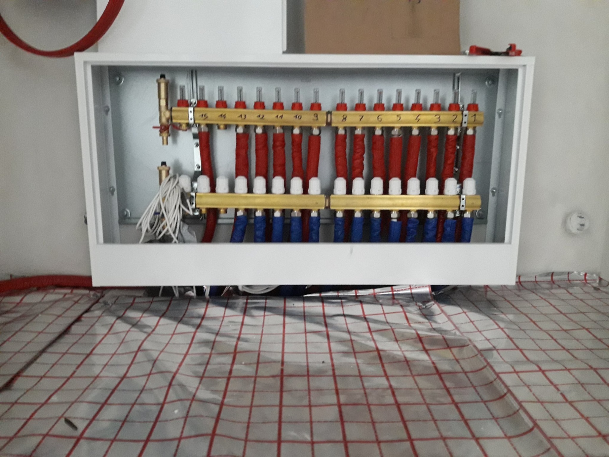 Rozdzielacz ogrzewania podłogowego w szafce, z czerwonymi i niebieskimi rurami, zamontowany na folii z siatką, widok z dołu.