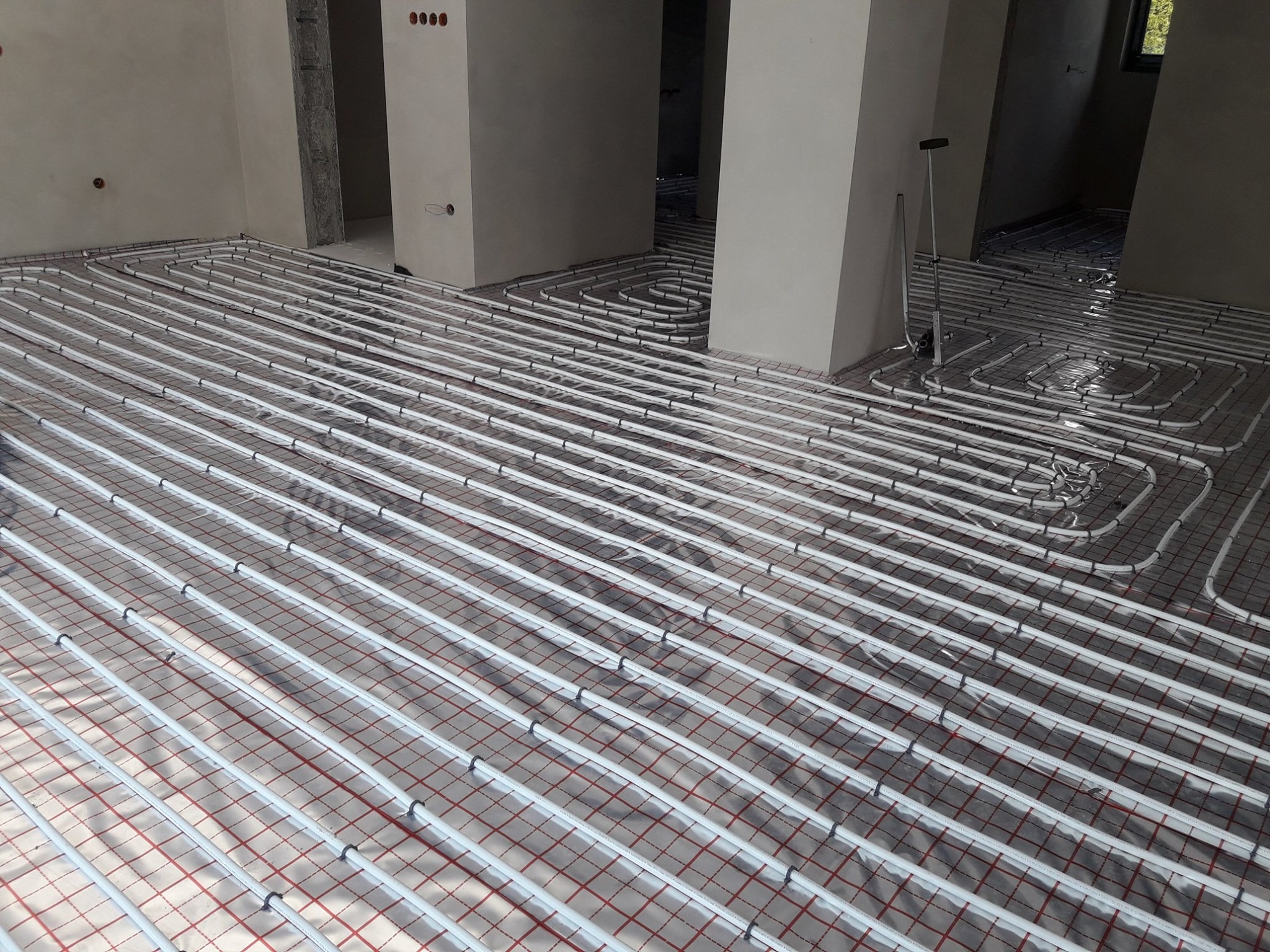 Rozłożone rury ogrzewania podłogowego na folii z rastrem w surowym wnętrzu, widoczny rozdzielacz i narzędzia instalacyjne.