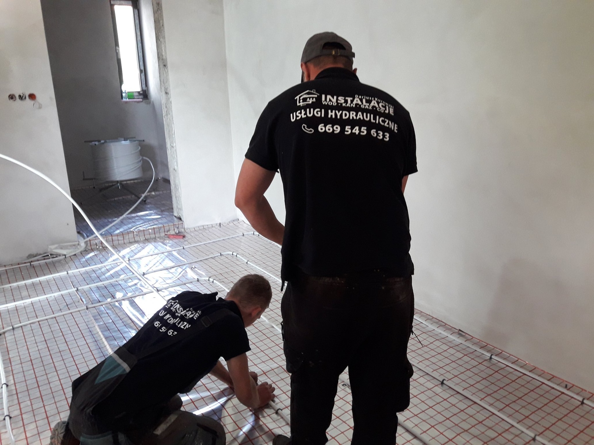 Dwóch instalatorów w trakcie montażu ogrzewania podłogowego, widoczna siatka montażowa i rury, jeden instalator w czarnej koszulce z logo firmy instalacyjnej, drugi w pozycji klęczącej przy podłodze.