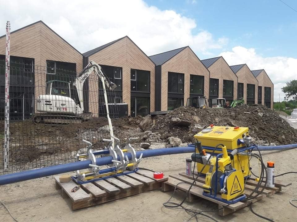 Zgrzewarka do rur z tworzyw sztucznych na palecie, na tle wykopu, minikoparki i szeregu budynków w trakcie budowy.