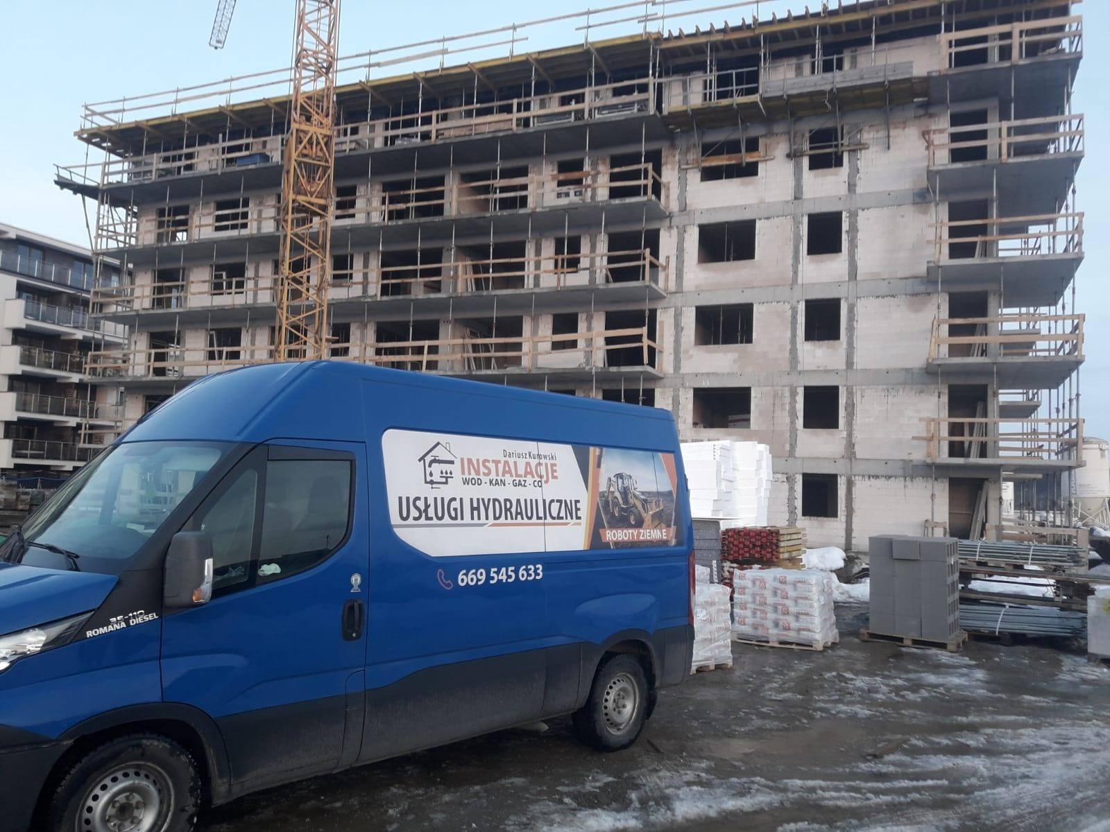 Niebieski van z logo firmy hydraulicznej na tle surowego budynku w trakcie budowy, widoczne materiały budowlane i żuraw.