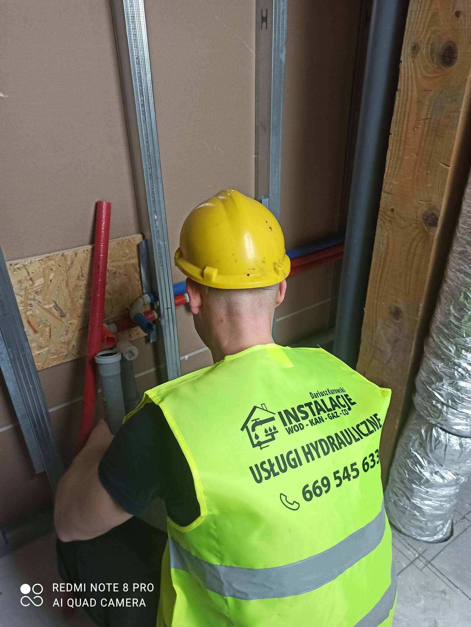 Hydraulik w żółtym kasku i odblaskowej kamizelce z logo firmy instalacyjnej, montuje rury instalacji wodnej wewnątrz budynku z widocznymi elementami konstrukcji metalowej i drewnianej.