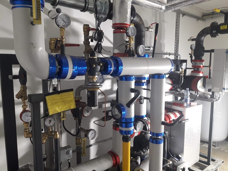 Rozbudowany system rur i zaworów w kotłowni, rury owinięte niebieską folią, liczne manometry i sterowniki, widoczny fragment pompy obiegowej i zbiornika wyrównawczego.