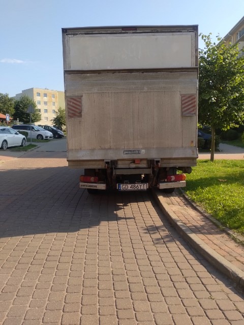 Tył brudnego samochodu dostawczego z gdańską rejestracją GD 486YT zaparkowanego na brukowanym parkingu w słoneczny dzień. Widoczne budynki w tle i drzewo po prawej stronie.