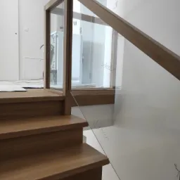 Fragment dębowych schodów z balustradą ze szkła, na której widać odręczne notatki, w tle białe ściany i drzwi.