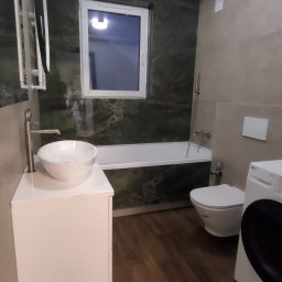 LEM-ART Remonty i Wykończenia Wnętrz Piotr Lemański - Nowoczesna łazienka po remoncie: umywalka na szafce, toaleta, wanna pod oknem i pralka. Ściany wykończone płytkami w odcieniach szarości i zieleni.