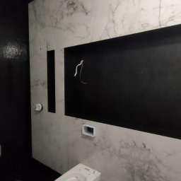 LEM-ART Remonty i Wykończenia Wnętrz Piotr Lemański - Nowoczesna łazienka w trakcie wykańczania: białe marmurowe płytki, czarne akcenty i wisząca toaleta. Widoczne przewody elektryczne w przygotowanej wnęce.