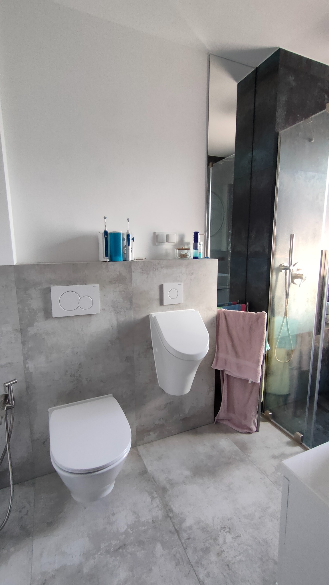 Nowoczesna łazienka w szarościach: podwieszana toaleta i bidet, płytki imitujące beton, minimalistyczna armatura, kabina prysznicowa z ciemnymi elementami.