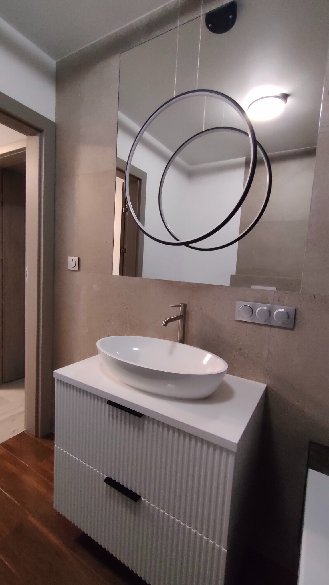 Nowoczesna łazienka z owalną umywalką nablatową, szafką z frezowanymi frontami i okrągłą lampą wiszącą odbijającą się w lustrze.