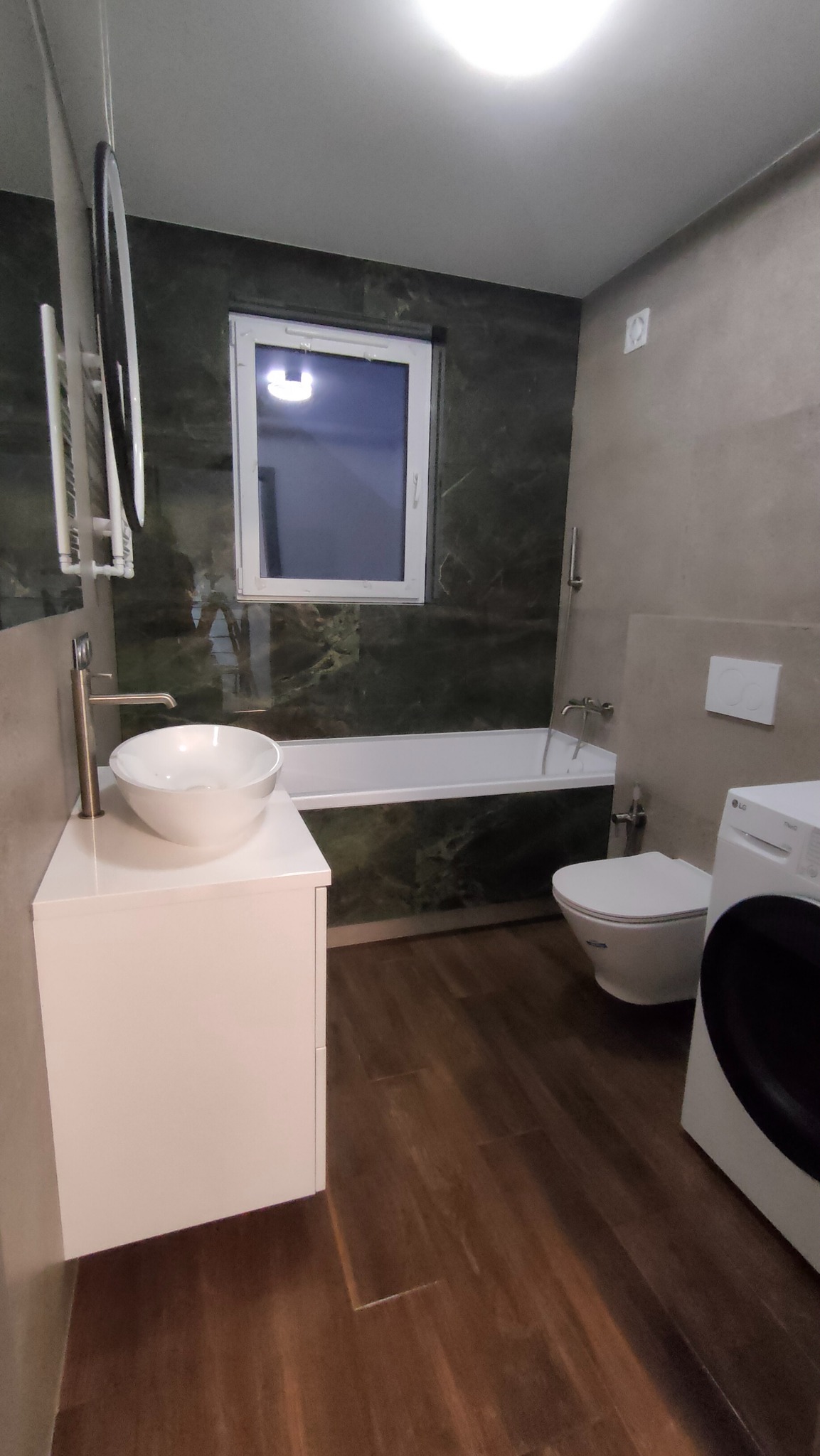 Nowoczesna łazienka po remoncie: umywalka na szafce, toaleta, wanna pod oknem i pralka. Ściany wykończone płytkami w odcieniach szarości i zieleni.