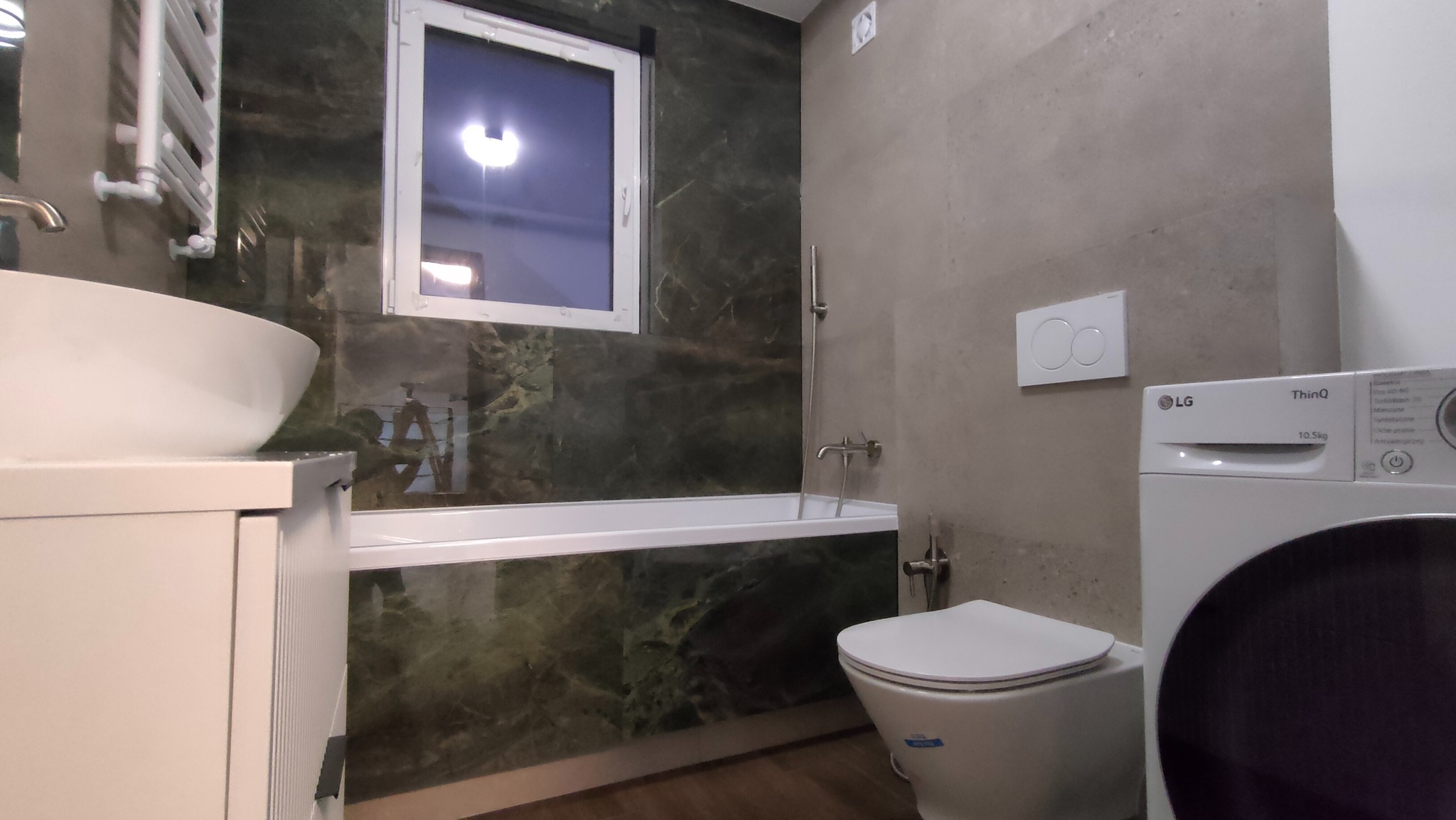 Nowoczesna łazienka z wanną obudowaną zielonym marmurem, umywalką nablatową i pralką LG. Minimalistyczny design, szare płytki, biała armatura i okno.