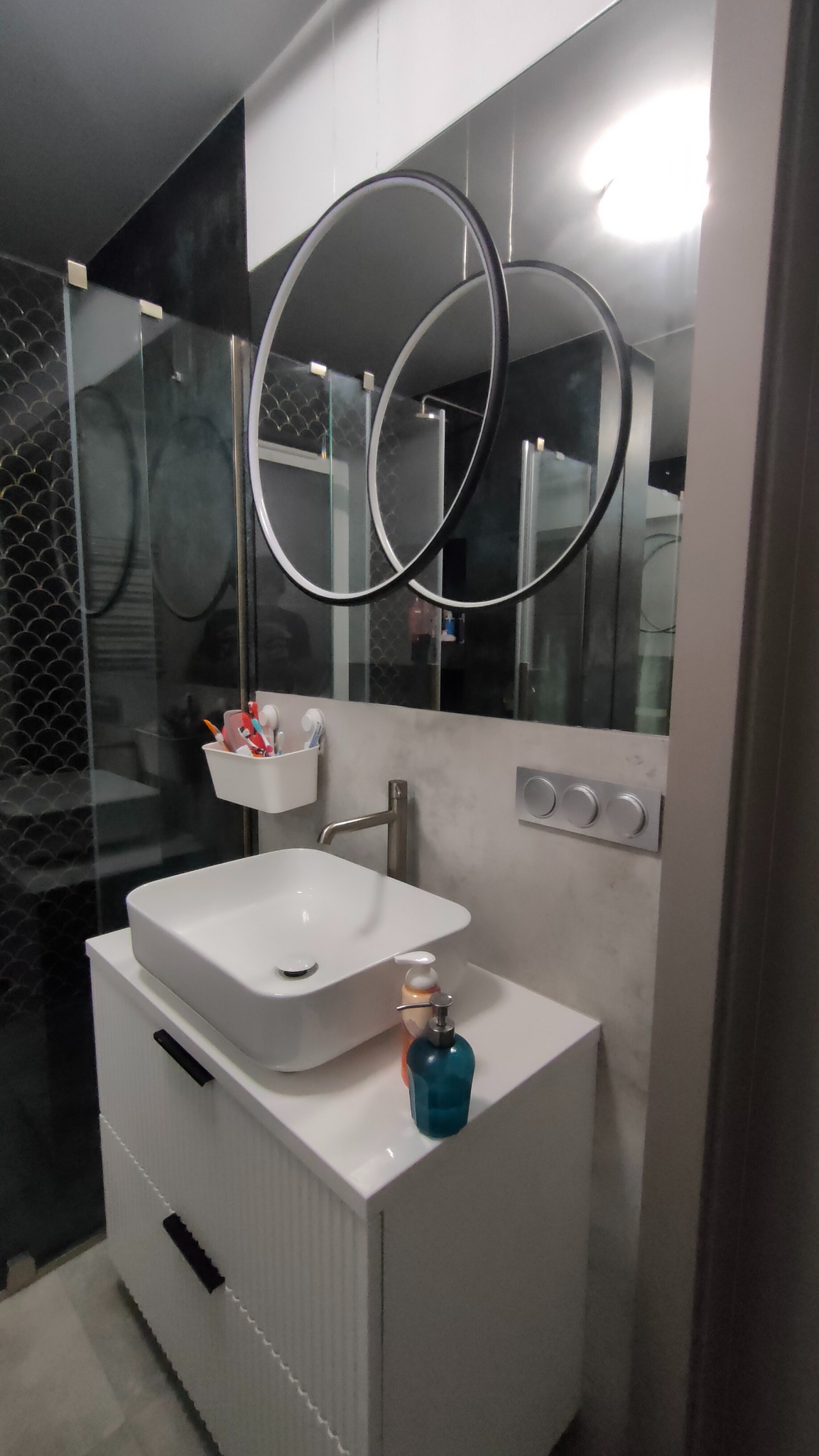 Nowoczesna łazienka z białą umywalką nablatową na szafce z frezowanymi frontami, okrągłe lustro z podświetleniem i czarne płytki na ścianie.