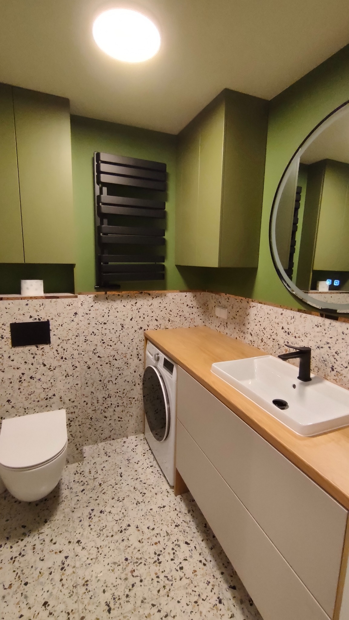 Nowoczesna łazienka w odcieniach zieleni z płytkami lastryko, czarnym grzejnikiem i okrągłym lustrem z podświetleniem LED. Umywalka nablatowa na szafce z drewnianym blatem.