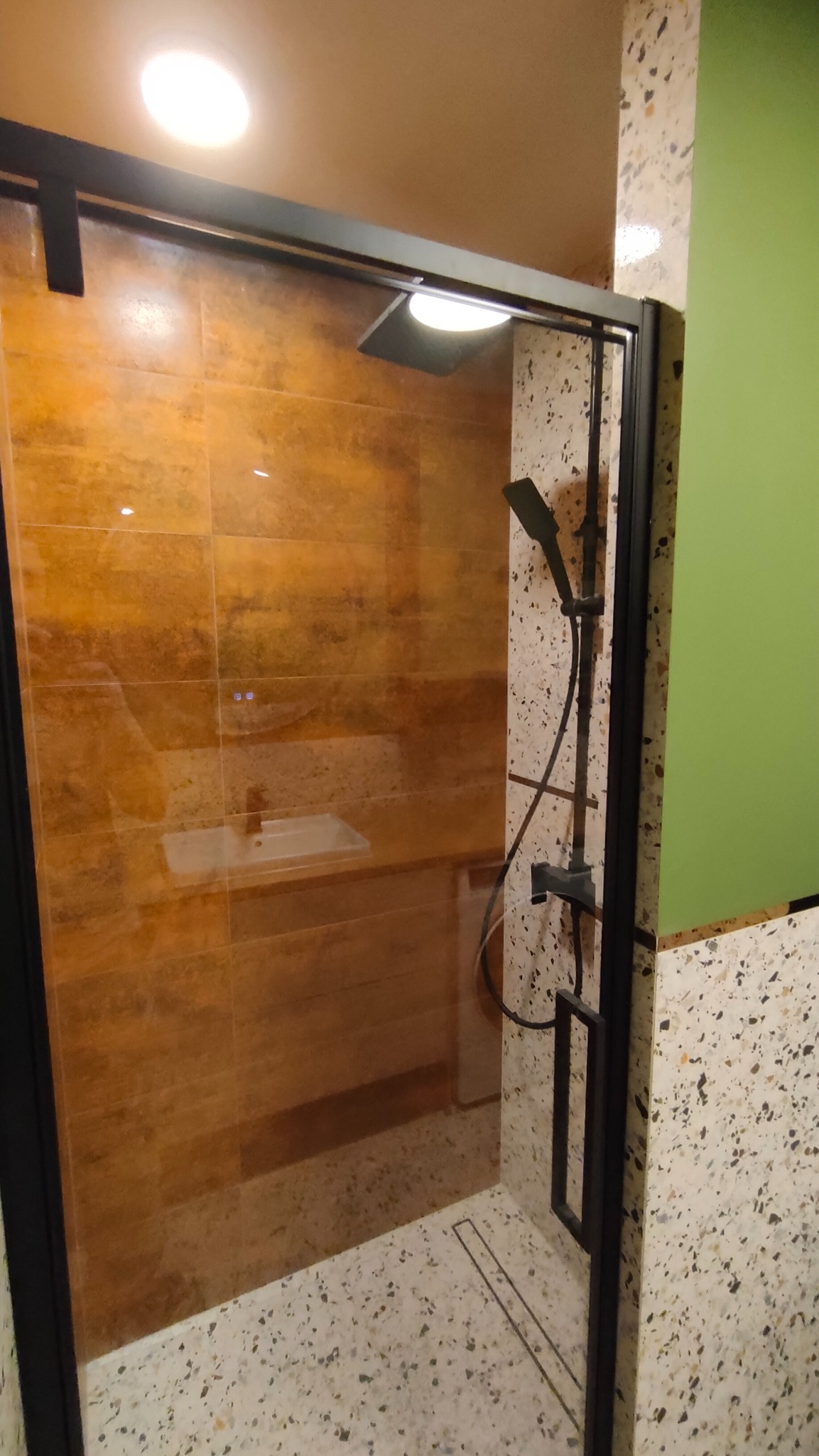 Kabina prysznicowa z czarnymi profilami, brązowe płytki imitujące kamień na ścianie i posadzka lastryko. Widoczna bateria prysznicowa i umywalka.