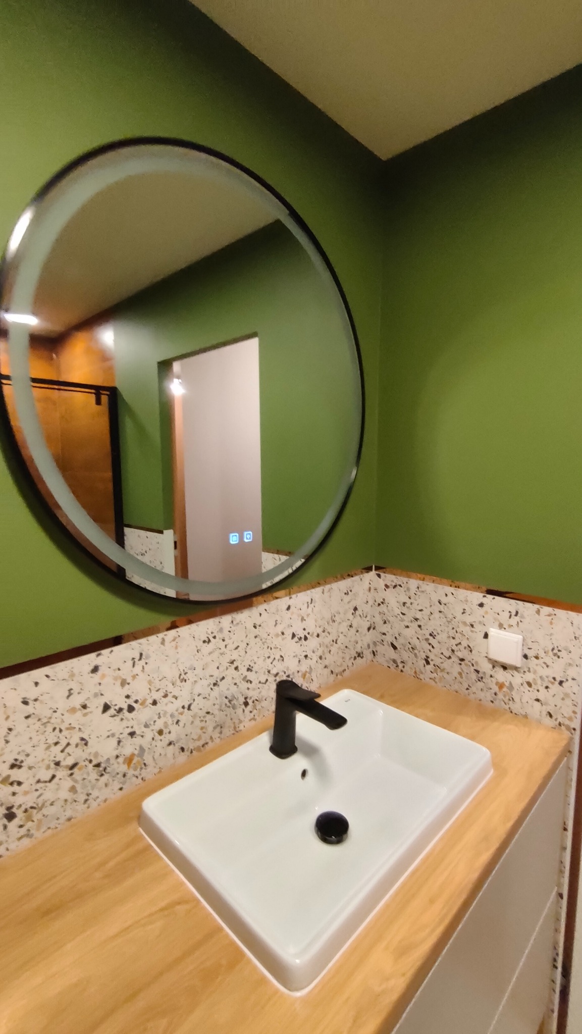 Nowoczesna łazienka z zielonymi ścianami, okrągłym lustrem z podświetleniem i umywalką nablatową z czarną armaturą. Blat drewnopodobny, ściana wykończona mozaiką.