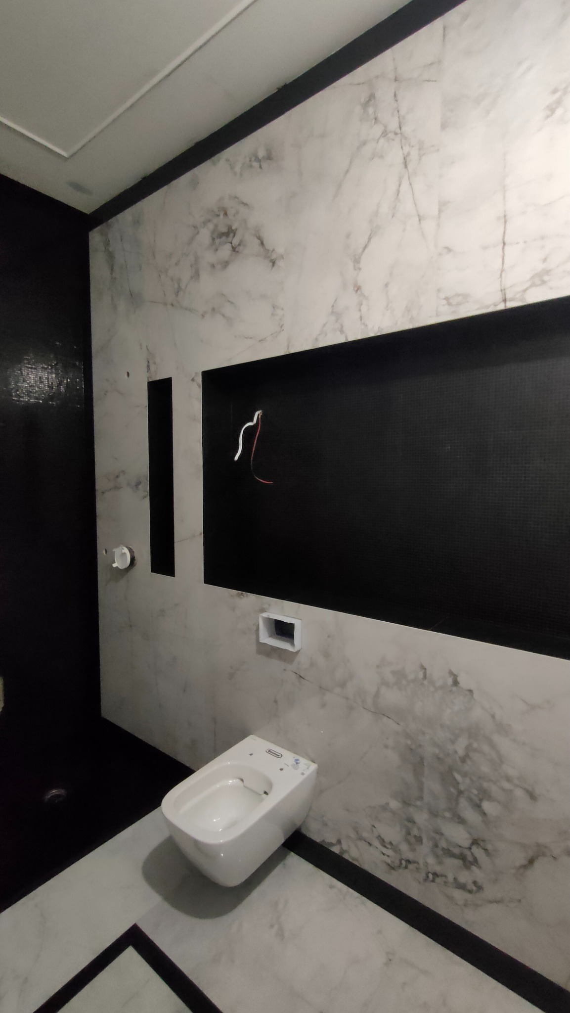 Nowoczesna łazienka w trakcie wykańczania: białe marmurowe płytki, czarne akcenty i wisząca toaleta. Widoczne przewody elektryczne w przygotowanej wnęce.