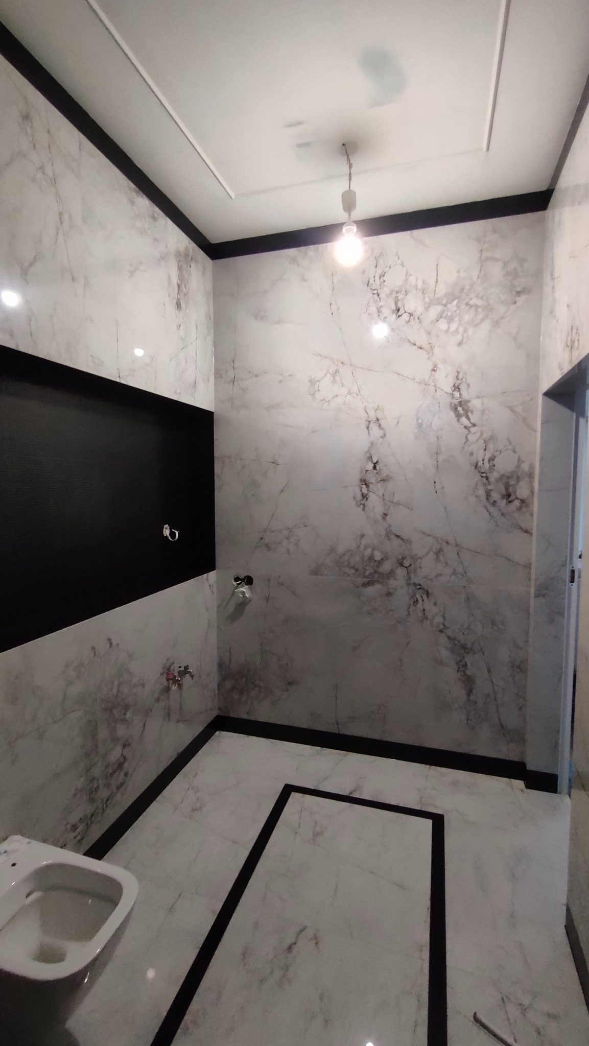 Wykończona łazienka z marmurowymi płytkami, czarnymi akcentami i wiszącą toaletą. Widoczne instalacje wodne i oświetlenie sufitowe.