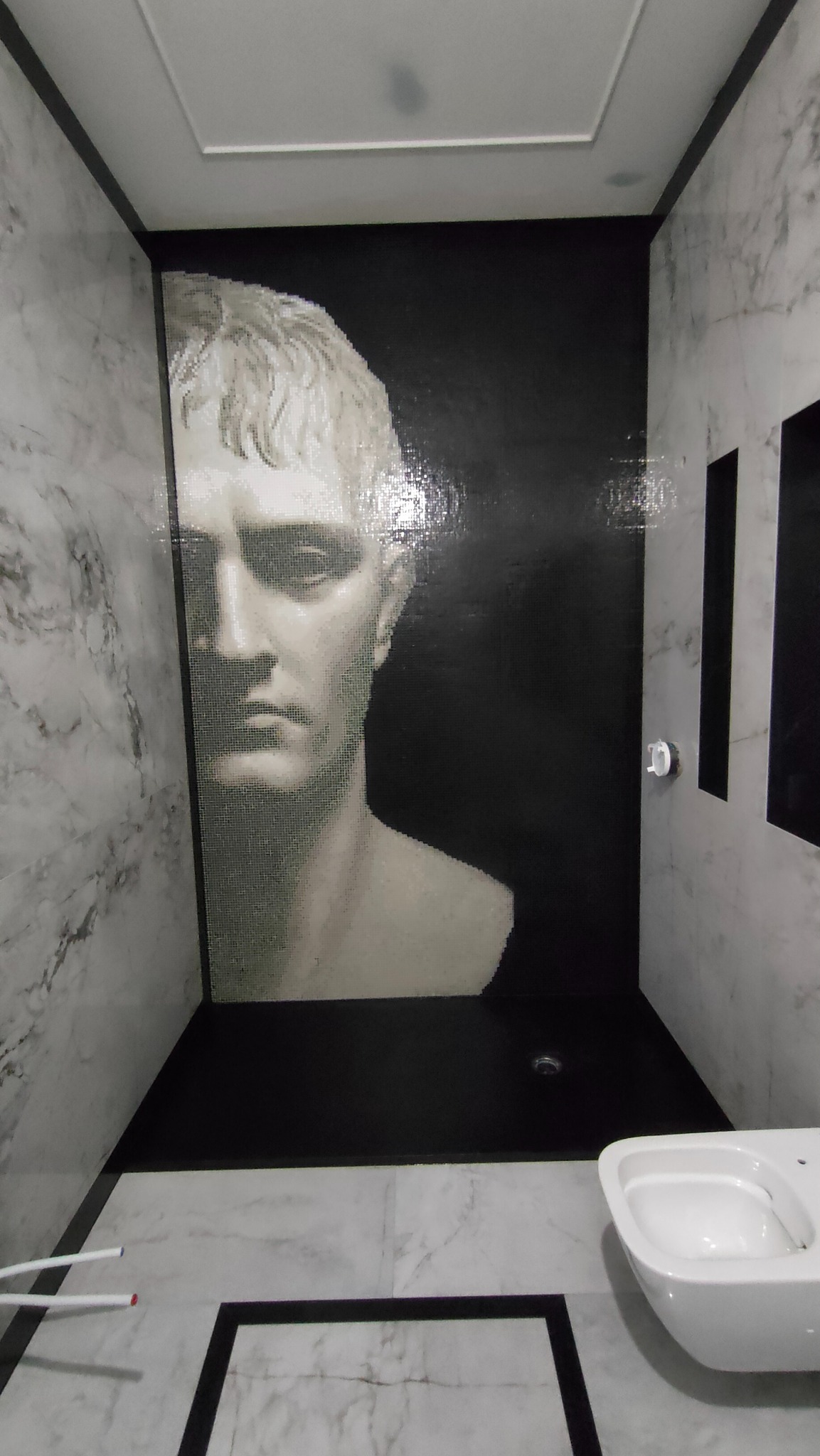 Nowoczesna łazienka z mozaiką portretu na czarnej ścianie, marmurowe płytki i wisząca toaleta. Minimalistyczny design z akcentem artystycznym.