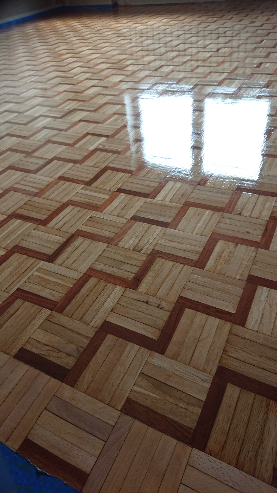 Lakierowany parkiet ułożony we wzór jodełki, z wyraźnym odbiciem światła w świeżej powłoce lakieru.