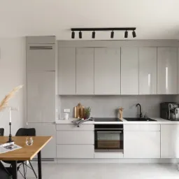 Nowoczesna, jasna kuchnia z szarymi, lakierowanymi szafkami, czarną armaturą i oświetleniem punktowym. Drewniany stół z dekoracjami w strefie jadalnej. Na blacie kuchennym deska do krojenia, zioła...