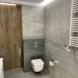 Nowoczesna łazienka z szarymi płytkami imitującymi marmur, podwieszana toaleta, drewnopodobna ściana prysznicowa i grzejnik drabinkowy