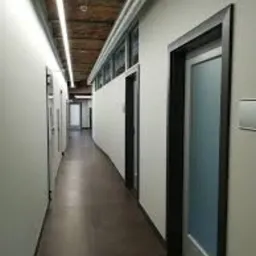 Korytarz biurowy z białymi ścianami, ciemną podłogą, oświetleniem LED pod sufitem i drzwiami z ciemnymi ościeżnicami. Widoczne fragmenty instalacji pod sufitem.