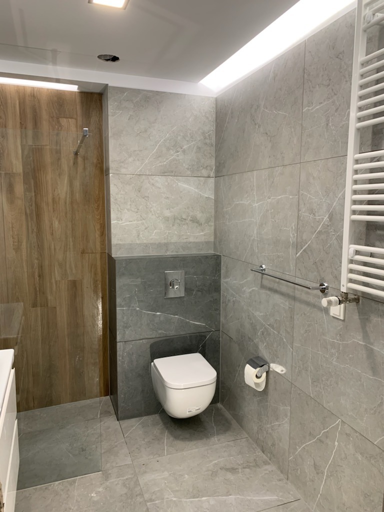 Nowoczesna łazienka z szarymi płytkami imitującymi marmur, podwieszana toaleta, drewnopodobna ściana prysznicowa i grzejnik drabinkowy