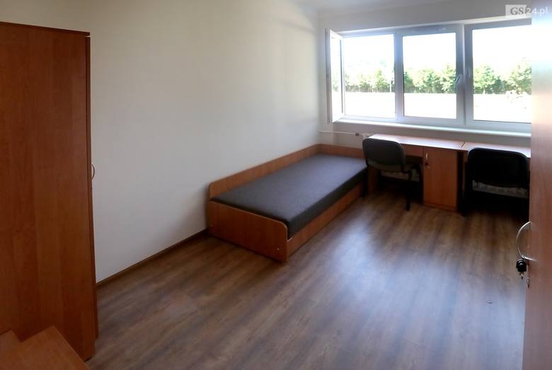 Wnętrze pokoju z drewnianą podłogą, pojedynczym łóżkiem z szarym materacem, biurkiem z dwoma krzesłami i szafą, widok z otwartych drzwi.